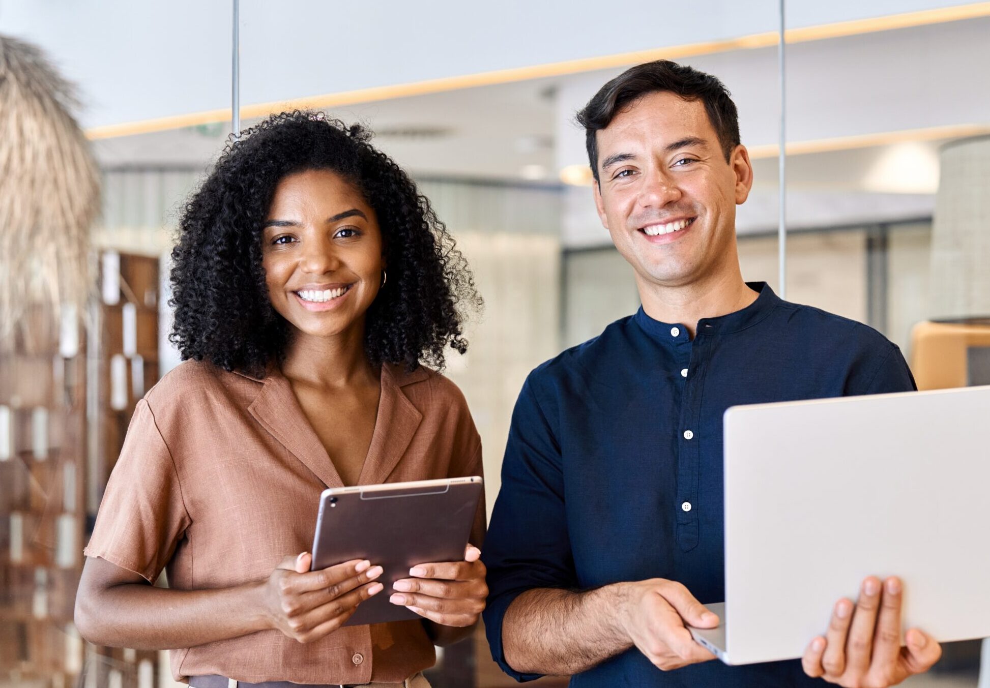 Two smiling diverse professionals standing in a modern office, holding a laptop and tablet, showcasing collaboration and innovation.